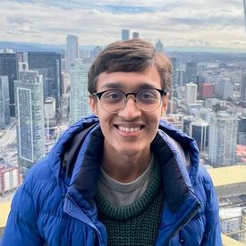A young man with brown hair and glasses smiles, high above a big city skyline.