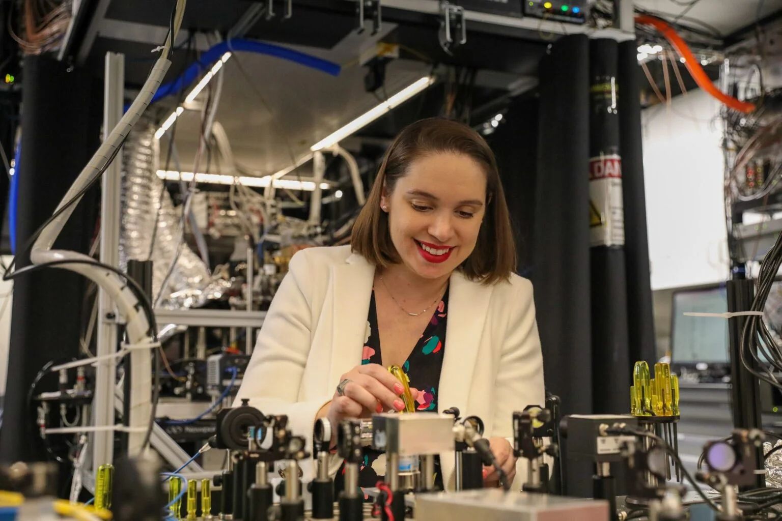 Crystal Noel smiles while adjusting complex laser equipment and optical components in a quantum engineering laboratory.