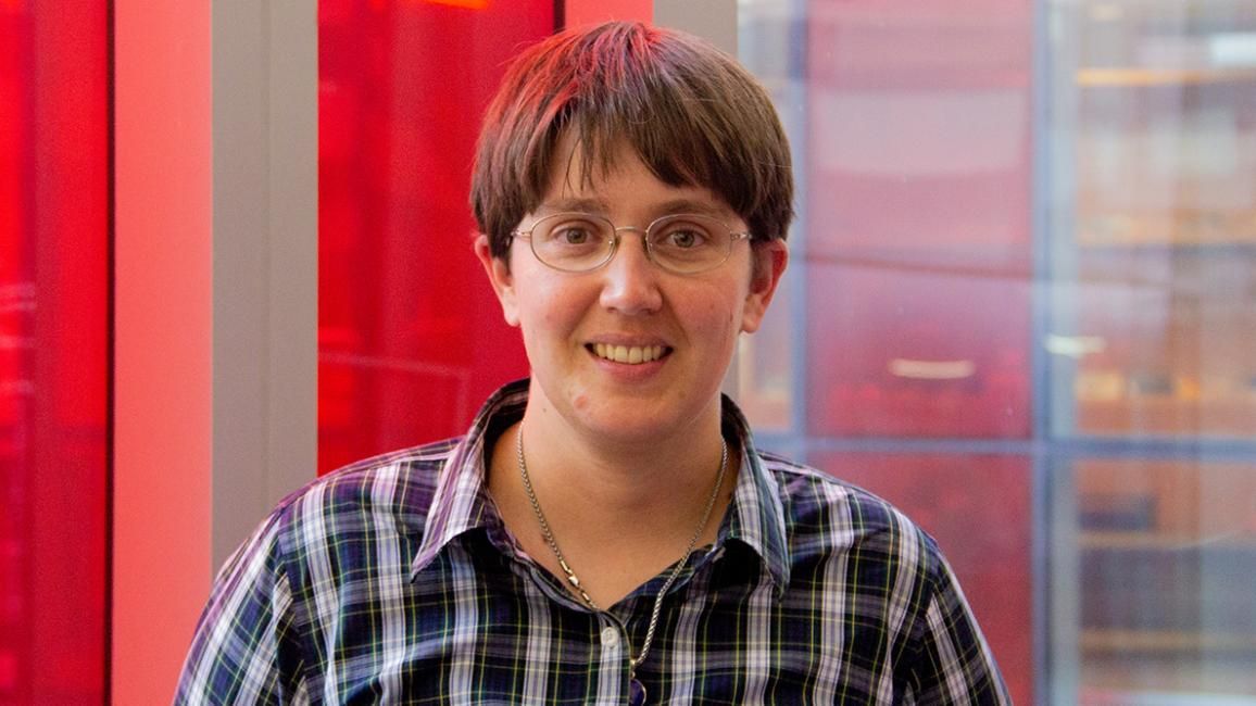 Alicia  Kollár wearing glasses and a blue and white plaid shirt in front of red and clear windows.