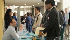 A young man speaks to another man seated at a desk at an event.