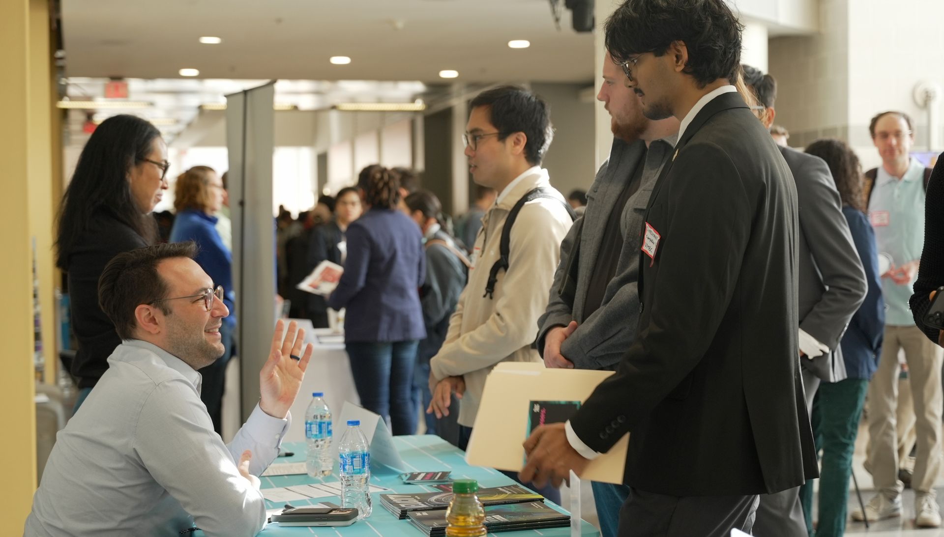 A young man speaks to another man seated at a desk at an event.