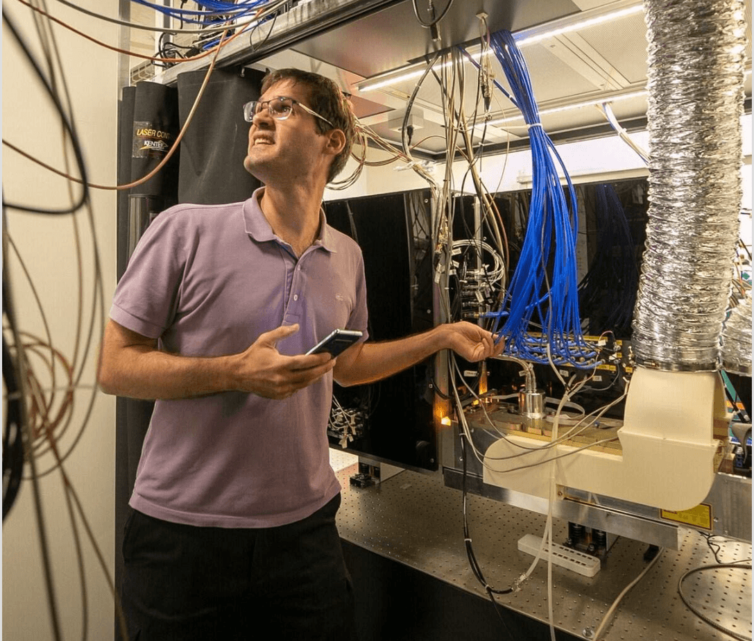 Marko Cetina wearing safety glasses and holding a phone while adjusting complex laboratory wiring and equipment.